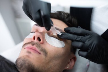 Erkek müşteri, modern bir güzellik salonunda kirpik uzatma uygulamasından geçiyor. Güzellik uzmanı, kusursuz bir görünüm için yapıştırıcı ve koruyucu pedler takıyor.