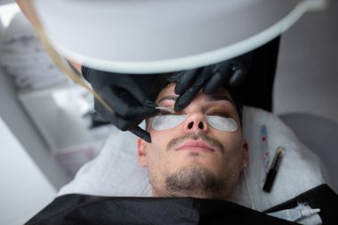 Erkek müşteri bir güzellik salonunda kirpik uzatma tedavisi görürken uzanıyor. Güzellik uzmanı yapıştırıcı sürüyor ve güzellik hizmetleri deneyimini vurguluyor.