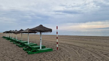 Sazlı şemsiyeli plaj salonları, sakin deniz ve bulutlu gökyüzüne bakan kumlu sahilde, ziyaretçiler için huzurlu bir kıyı inzivasını çağrıştırıyor.