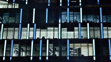Geceleri şehirdeki modern ofis binası. Bir şirket binasının aydınlatılmış ofislerine bakın. Cam ve çelikten yapılmış çok katlı binanın penceresinde yanıp sönen ışık. Geceleri uzun pozlama