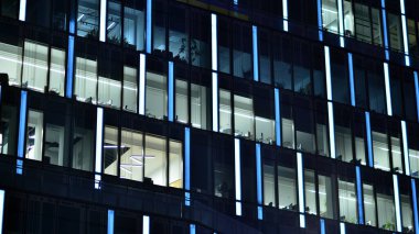 Geceleri şehirdeki modern ofis binası. Bir şirket binasının aydınlatılmış ofislerine bakın. Cam ve çelikten yapılmış çok katlı binanın penceresinde yanıp sönen ışık. Geceleri uzun pozlama