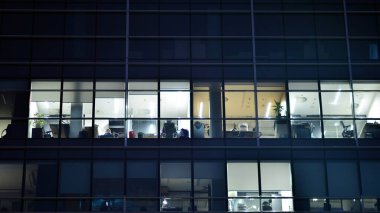 Geceleri şehirdeki modern ofis binası. Bir şirket binasının aydınlatılmış ofislerine bakın. Cam ve çelikten yapılmış çok katlı binanın penceresinde yanıp sönen ışık. Geceleri uzun pozlama