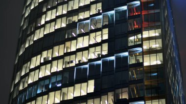 Geceleri şehirdeki modern ofis binası. Bir şirket binasının aydınlatılmış ofislerine bakın. Cam ve çelikten yapılmış çok katlı binanın penceresinde yanıp sönen ışık. Geceleri uzun pozlama