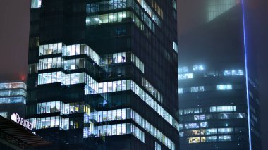 Geceleri şehirdeki modern ofis binası. Bir şirket binasının aydınlatılmış ofislerine bakın. Cam ve çelikten yapılmış çok katlı binanın penceresinde yanıp sönen ışık. Geceleri uzun pozlama