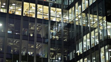 Geceleri şehirdeki modern ofis binası. Bir şirket binasının aydınlatılmış ofislerine bakın. Cam ve çelikten yapılmış çok katlı binanın penceresinde yanıp sönen ışık. Geceleri uzun pozlama