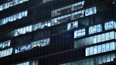 Geceleri şehirdeki modern ofis binası. Bir şirket binasının aydınlatılmış ofislerine bakın. Cam ve çelikten yapılmış çok katlı binanın penceresinde yanıp sönen ışık. Geceleri uzun pozlama