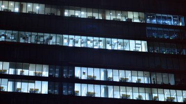 Geceleri şehirdeki modern ofis binası. Bir şirket binasının aydınlatılmış ofislerine bakın. Cam ve çelikten yapılmış çok katlı binanın penceresinde yanıp sönen ışık. Geceleri uzun pozlama