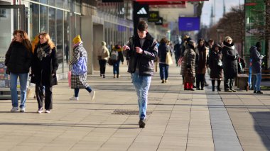 Varşova, Polonya. 8 Mart 2023. Şehir merkezindeki işlek bir caddenin arka planında parlayan güneş ışığıyla şehir kaldırımında yürüyen bir kalabalık..