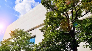 Mavi gökyüzüne karşı modern beyaz beton bina duvarları. Çevre mimarisi. Yeşil ağaçlar ve beton ofis binası. Doğanın ve modernliğin ahengi.