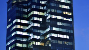 Geceleri modern bir gökdelenin cam cephesi. Şehirde modern cam ofis. Geceleri modern ofis binasında parlayan büyük pencereler. Pencere sıralarında ışık parlıyor.