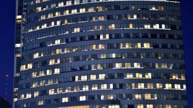 Geceleri modern bir gökdelenin cam cephesi. Şehirde modern cam ofis. Geceleri modern ofis binasında parlayan büyük pencereler. Pencere sıralarında ışık parlıyor.