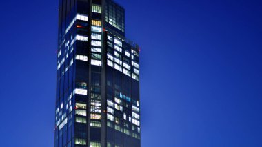 Geceleri modern bir gökdelenin cam cephesi. Şehirde modern cam ofis. Geceleri modern ofis binasında parlayan büyük pencereler. Pencere sıralarında ışık parlıyor.