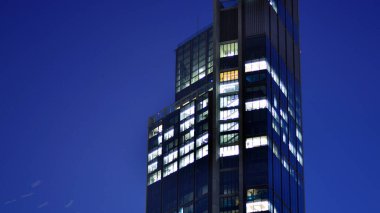 Geceleri modern bir gökdelenin cam cephesi. Şehirde modern cam ofis. Geceleri modern ofis binasında parlayan büyük pencereler. Pencere sıralarında ışık parlıyor.