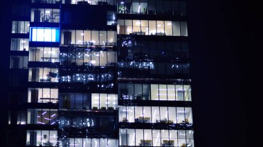 Geceleri modern bir gökdelenin cam cephesi. Şehirde modern cam ofis. Geceleri modern ofis binasında parlayan büyük pencereler. Pencere sıralarında ışık parlıyor.