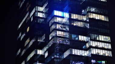 Geceleri modern bir gökdelenin cam cephesi. Şehirde modern cam ofis. Geceleri modern ofis binasında parlayan büyük pencereler. Pencere sıralarında ışık parlıyor.