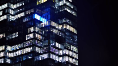 Geceleri modern bir gökdelenin cam cephesi. Şehirde modern cam ofis. Geceleri modern ofis binasında parlayan büyük pencereler. Pencere sıralarında ışık parlıyor.