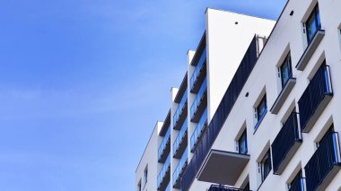 Beyaz, modern, yüksek katlı apartman binası, mavi gökyüzüne karşı dik dur, parlak, açısal tasarımlarla yenilikçi şehir mimarisini örnek al..