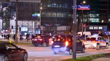 Varşova, Polonya. 21 Kasım 2024. Geceleri sokakta arabaların sokak fotoğrafçılığı. Modern binanın arka planındaki ışık izleri.