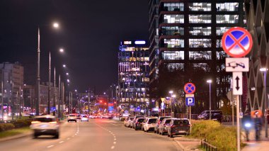 Varşova, Polonya. 21 Kasım 2024. Geceleri sokakta arabaların sokak fotoğrafçılığı. Modern binanın arka planındaki ışık izleri.