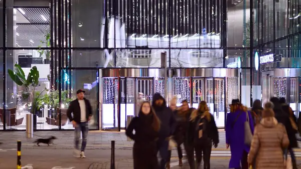 Varşova, Polonya. 21 Kasım 2024. Geceleri sokaktaki insanların sokak fotoğrafçılığı. Modern binanın arka planındaki yaya siluetleri. İnsanların hareket bulanıklığı. Şehir hayatı kavramı.