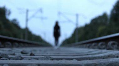 Demiryolu raylarında yürüyen bir kadının bulanık silueti. Ön plandaki taşlara keskin bir dikkatle odaklanmış..