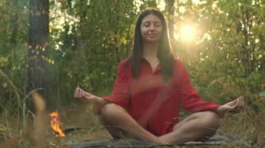 Huzurlu bir ormanda meditasyon yapan bir kadın, küçük bir kamp ateşinin yanında bacak bacak üstüne atarak oturur güneş ağaçların arasından batarken, huzur ve doğa ile kucaklaşır..