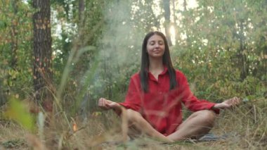 Kırmızı gömlekli bir kadın, sakin bir ormanda meditasyon yapıyor, ağaçların arasından güneş parlıyor, küçük bir kamp ateşinin yanında, barışçıl bir inziva sırasında yan yana oturuyor..