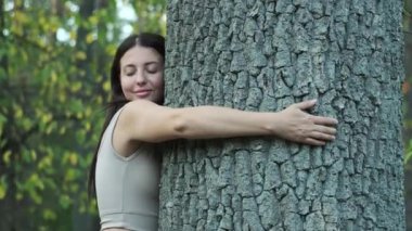 Genç bir kadın gözleri kapalı gülümsüyor, ormanın içinde bir ağaca sarılıyor..