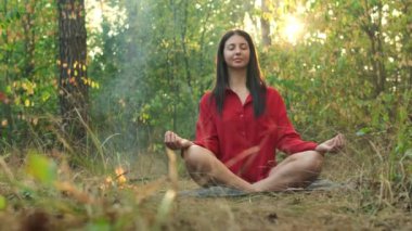 Huzurlu bir ormanda meditasyon yapan bir kadın, küçük bir kamp ateşinin yanında bacak bacak üstüne atarak oturur güneş ağaçların arasından batarken, huzur ve doğa ile kucaklaşır..