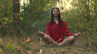 Kırmızı gömlekli bir kadın, sakin bir ormanda meditasyon yapıyor, ağaçların arasından güneş parlıyor, küçük bir kamp ateşinin yanında, barışçıl bir inziva sırasında yan yana oturuyor..