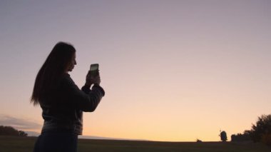 Akıllı telefonuyla güzel bir akşam manzarası yakalayan genç bir kadın gün batımına karşı koyuyor..