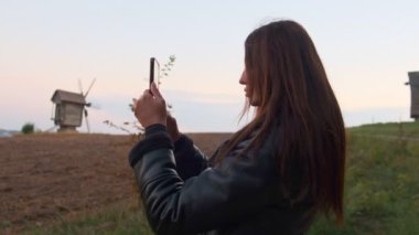 Bir turist, ufukta tarihi ahşap yel değirmenleri olan bir araziyi çerçeveleyerek akıllı telefon kamerasını en iyi çekim için ayarlıyor..