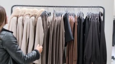 Bir kadın elbise askısı üzerindeki kışlıkları inceliyor, şık bir butikte kumaş ve stili değerlendirmek için elbiselere dokunuyor.