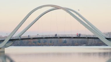 İnsanlar günbatımında modern bir köprüde huzur içinde yürüyor, kentsel tasarımla doğal çevreyi harmanlıyor..