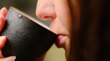 Siyah bir seramik bardaktan çay yudumlayan dudakların yakın görüntüsü, sakinliği ve farkındalığı yansıtır..