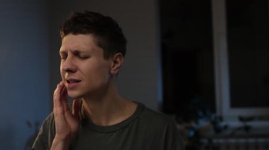 Diş sorunları. Yanağa dokunan genç adam, korkunç bir ifadeyle gözlerini kapatan diş ağrısı, hassas dişler, çürükler. Evdeki beyaz adam oturma odasında..