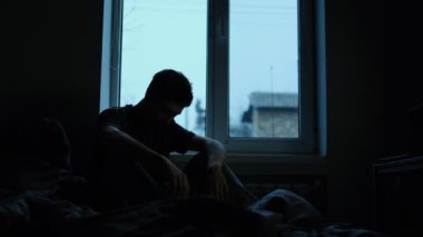 Derin duygusal sıkıntı içindeki bir pencerenin yanındaki yatakta oturan genç bir adamın silüeti. Yüksek kalite 4k görüntü