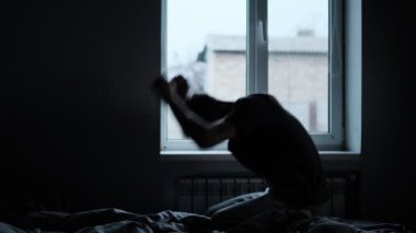 Bir adamın silueti pencerenin yanındaki bir yatağa oturur, umutsuzca yatağa vurur, duygusal karmaşayı yansıtır..