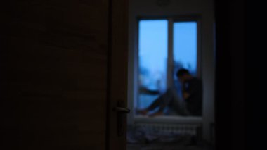 Bir adam karanlık bir odada pencere eşiğinde oturur, açık bir kapıdan bakar, duygusal yalnızlığı ve üzüntüyü ifade eder..