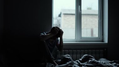 Derin duygusal sıkıntı içindeki bir pencerenin yanındaki yatakta oturan genç bir adamın silüeti. Yüksek kalite 4k görüntü