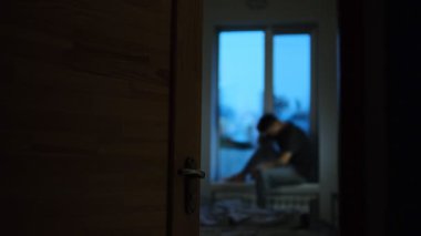 Pencerenin pervazında oturan bir adamın bulanık görüntüsü, açık bir kapıdan görülüyor, loş bir odada hüzün ve yalnızlığı iletiyor..