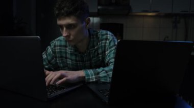Genç adam gece geç saatlere kadar iki dizüstü bilgisayarla çalışıyor, odaklanmış ve loş bir odada yoğunlaşmış. Çift ekran vurgulama teknolojisi ve çoklu görev.