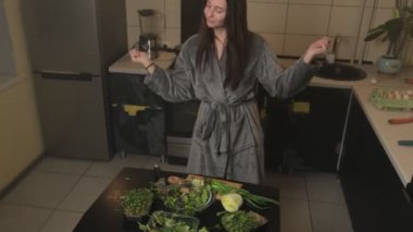 Cüppeli neşeli genç bir kadın vejetaryen yemeği hazırlıyor, mutfakta dans ederken salata kabına sebze atıyor..