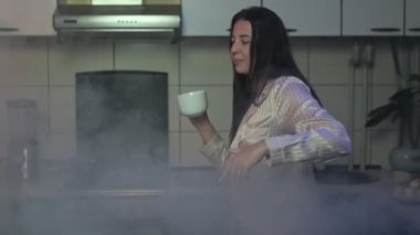 İpek pijamalı bir kadın neşeli bir şekilde dans ediyor dumanlı bir mutfakta çay ya da kahve içiyor, coşkulu ve kaygısız bir enerji ile parti havası yaratıyor.