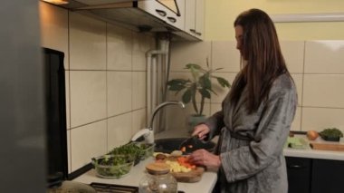 Yumuşak gri bornozlu bir kadın sıcak, ev yapımı bir mutfakta taze sebzeleri ahşap bir kesme tahtasıyla dikkatlice doğruyor ve ev yapımı yemek için malzemeler hazırlıyor..