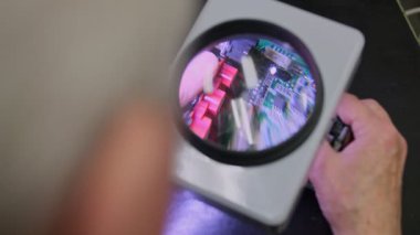 Elektronik devre kartının yakın çekim görüntüsü büyük bir büyüteçle büyütülmüş, teknisyenlerin elleri hassasiyet ve kalite kontrol bileşenlerini dikkatle inceliyor.
