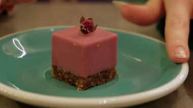 Kurumuş gül goncasıyla kaplanmış kremalı pembe kare bir tatlı, çıtır fındık kabuğunun üzerinde oturuyor, turkuaz bir tabakta güzelce sunuluyor.