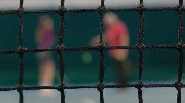 Bir tenis koçu bir acemiyi eğitir. Ön planda, top ona çarptığında ağ sallanıyor. Arka planda, bu sporda çalışmakla ve yeteneklerini geliştirmekle meşgul olan bulanık insanlar var.