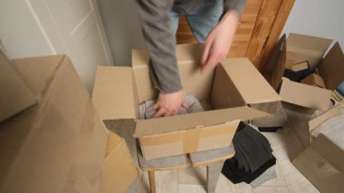 Bir adam taşınmaya hazırlanırken katlanmış giysileri açık bir karton kutuya yerleştirir. Etrafında bir sürü kutu, dağınık giysiler ve dağınık bir odada ambalaj malzemeleri var..