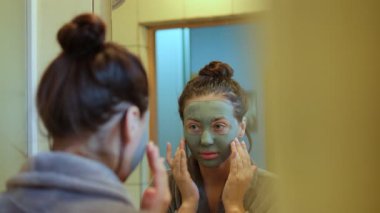 Yeşil kilden maskeli genç bir kadın aynada tenini inceliyor. Güzelliğinin ve cilt bakımının bir parçası olarak nazikçe yüzüne dokunuyor..